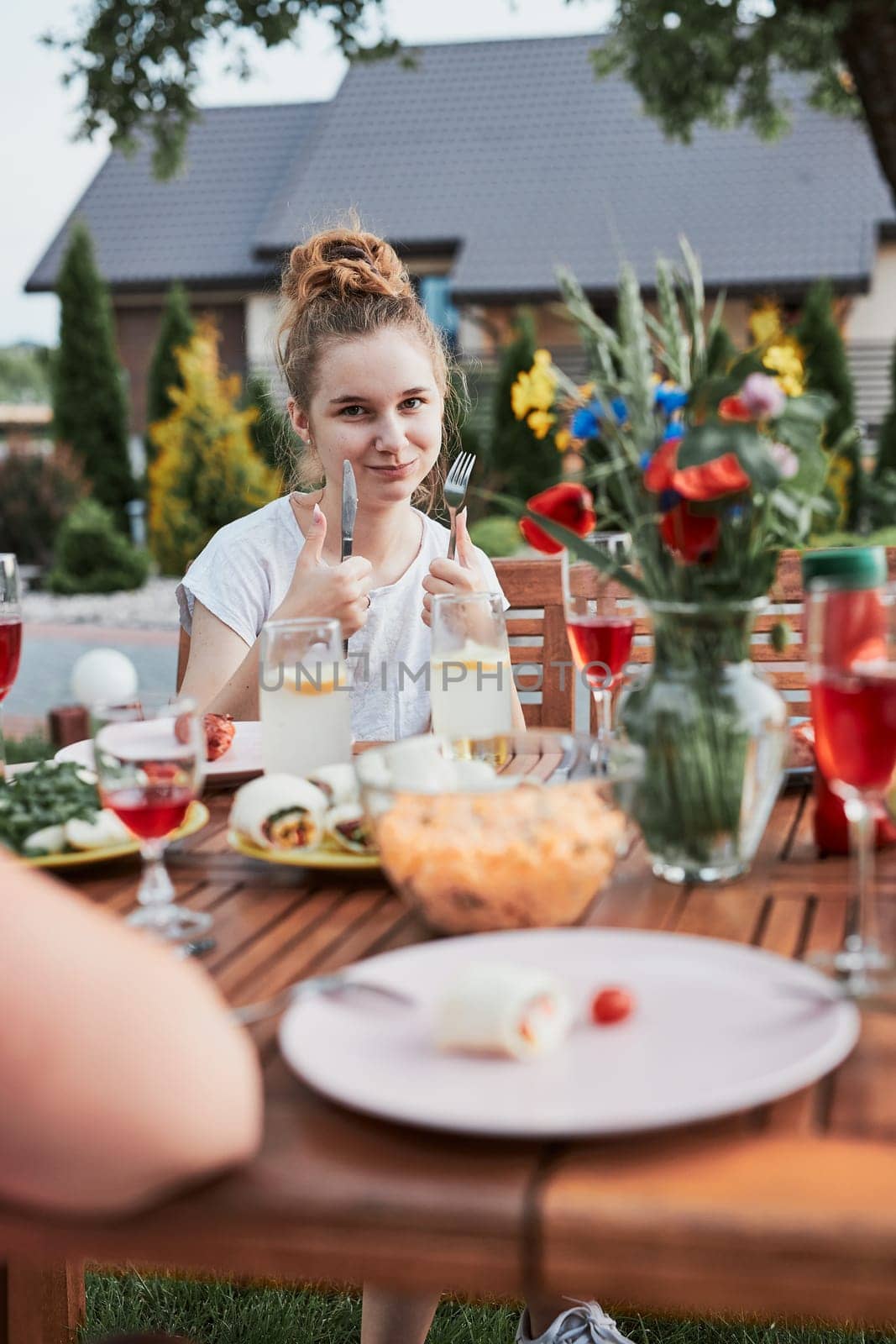 Young woman having a meal from grill during summer family picnic outdoor dinner in a home garden. Girl enjoying her tasty meal showing okay gesture. Close up of table with food and dishes