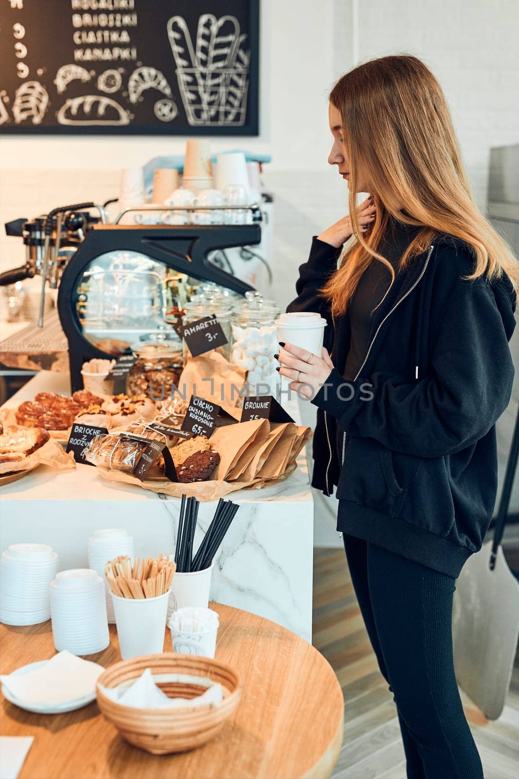 Woman holding cup with coffee looking at pastry, buns, cakes and cookies and waiting for the order. Girl buying a sweet food and hot drink to go. Young woman having a break doing shopping in a coffee shop