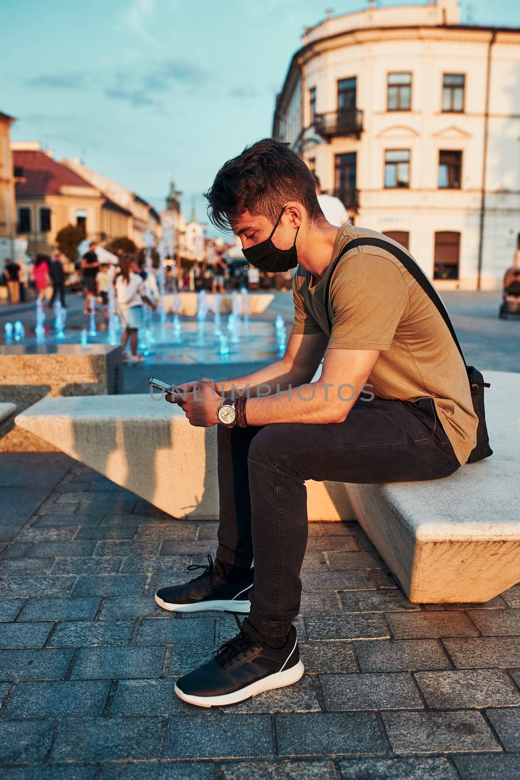 Young man sitting in the city center in the evening, using smartphone, wearing the face mask to avoid virus infection and to prevent the spread of disease in time of coronavirus