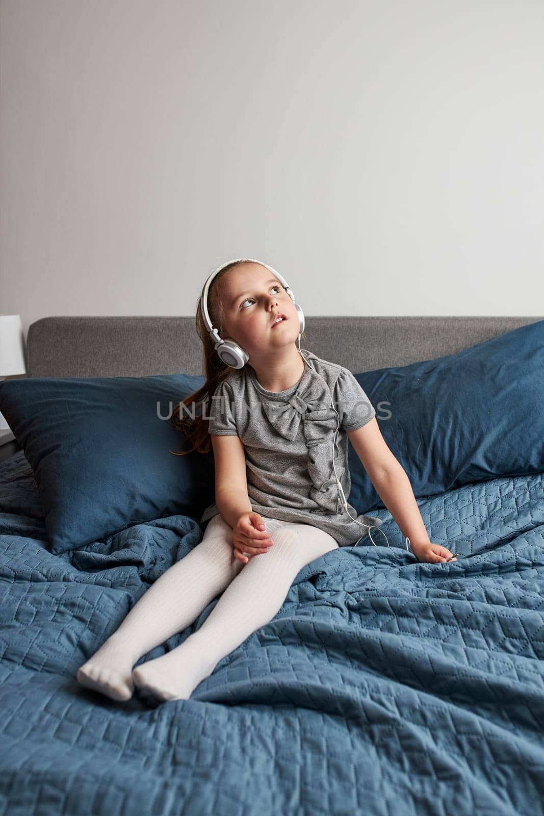 Little girl singing holding headphones cord imitating herself a real singer. Child having fun jumping dancing listening to music on bed in bedroom at home