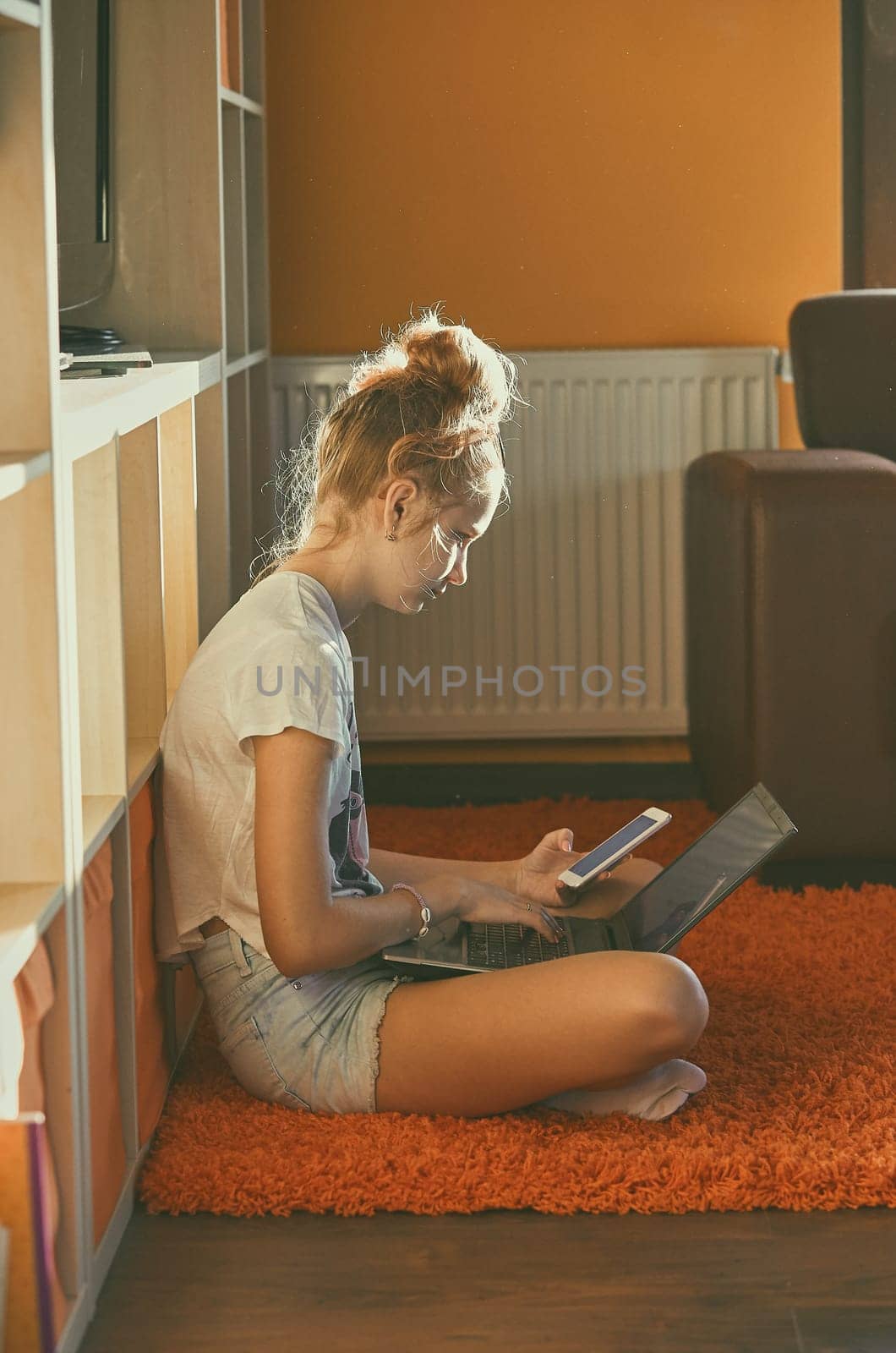 Young woman using portable computer and mobile phone, sitting on a floor, learning online at home. Candid people, real moments, authentic situations