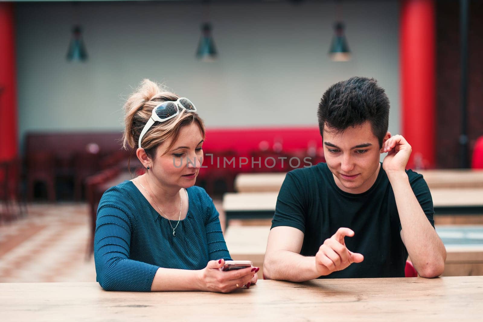 Woman and young man talking together and using mobile phones sitting by a table in cafe