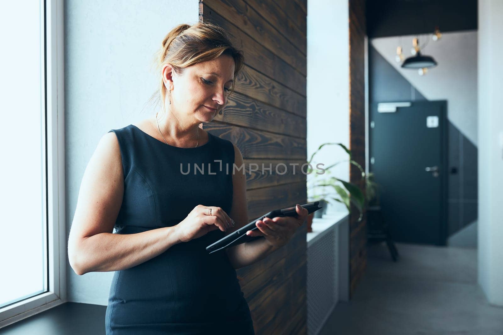 Businesswoman working on tablet in office. Mature woman using touch pad computer standing by window in modern interior. Female manager focused on work holding digital device. Using technology. Open office. Copy space right