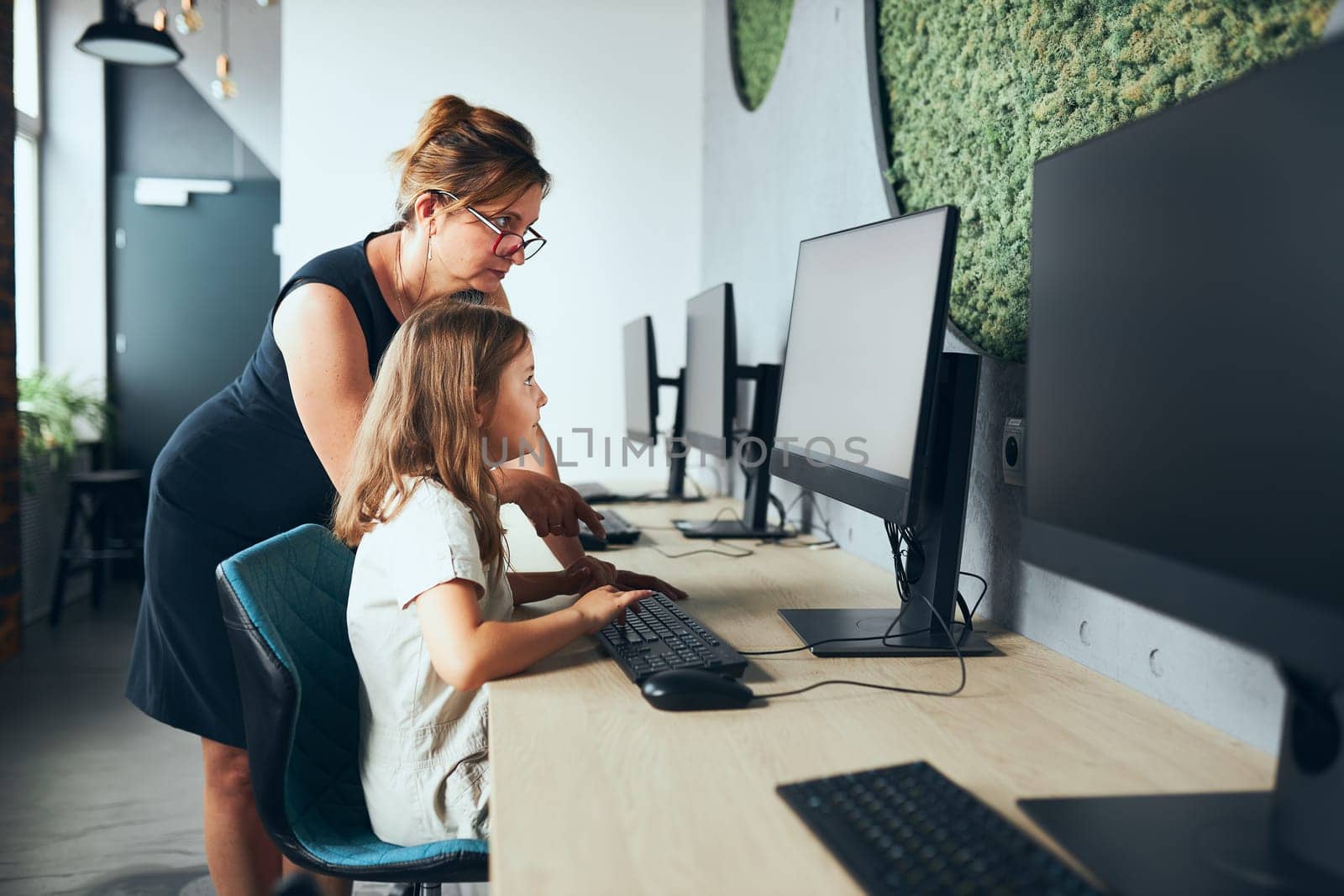 Computer class at school. Teacher assisting schoolgirl while class at primary school. Child learning computer on elementary computer science lesson. Teacher helping to find task solution. Giving clues. Smart little girl learning online at school. Back to school
