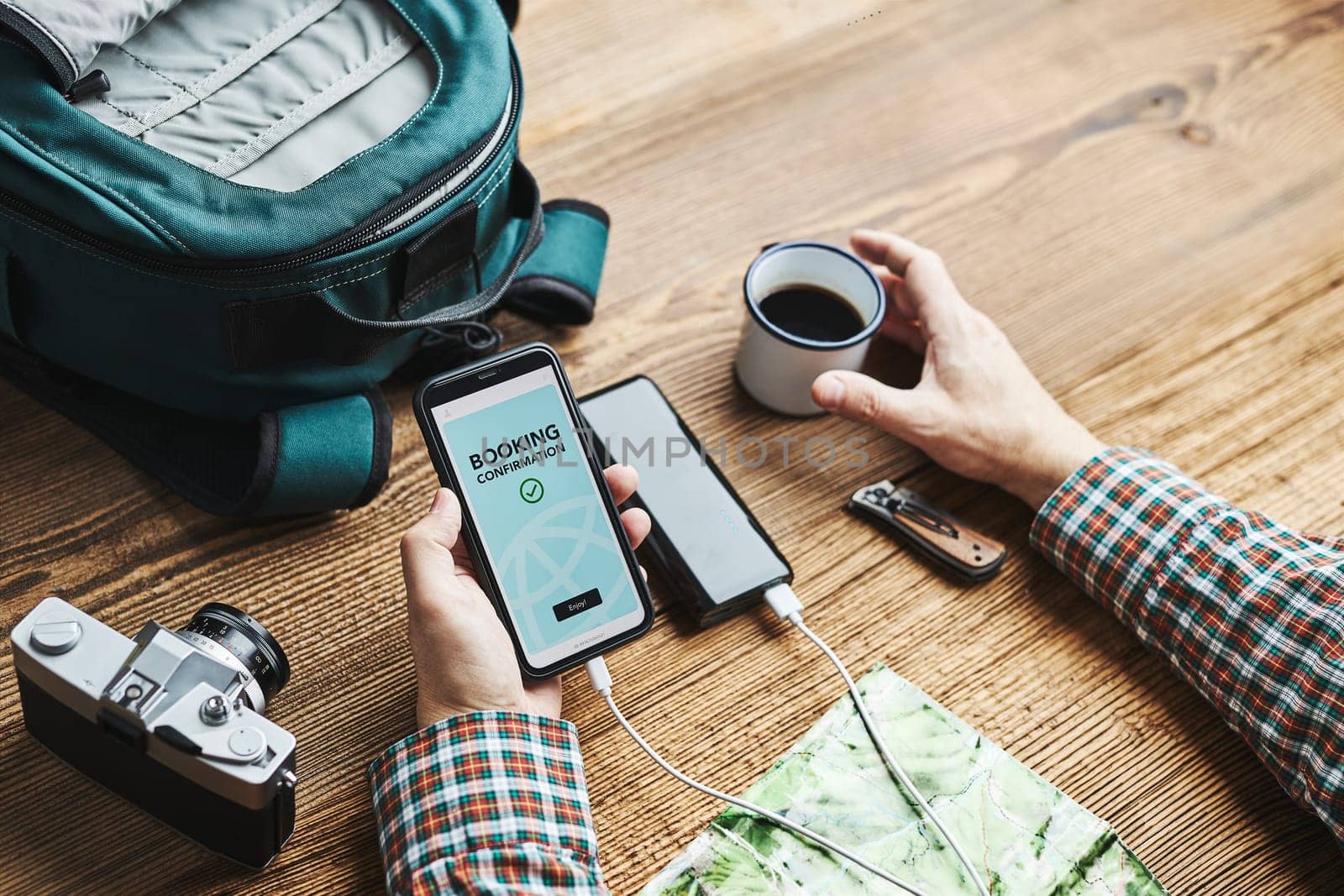 Man planning next vacation trip. Booking hotel online using smartphone. Ordering service. Charging mobile phone with power bank. Using technology while travelling. Man sitting at wooden table with camera and backpack drinking coffee. Travel plan concept