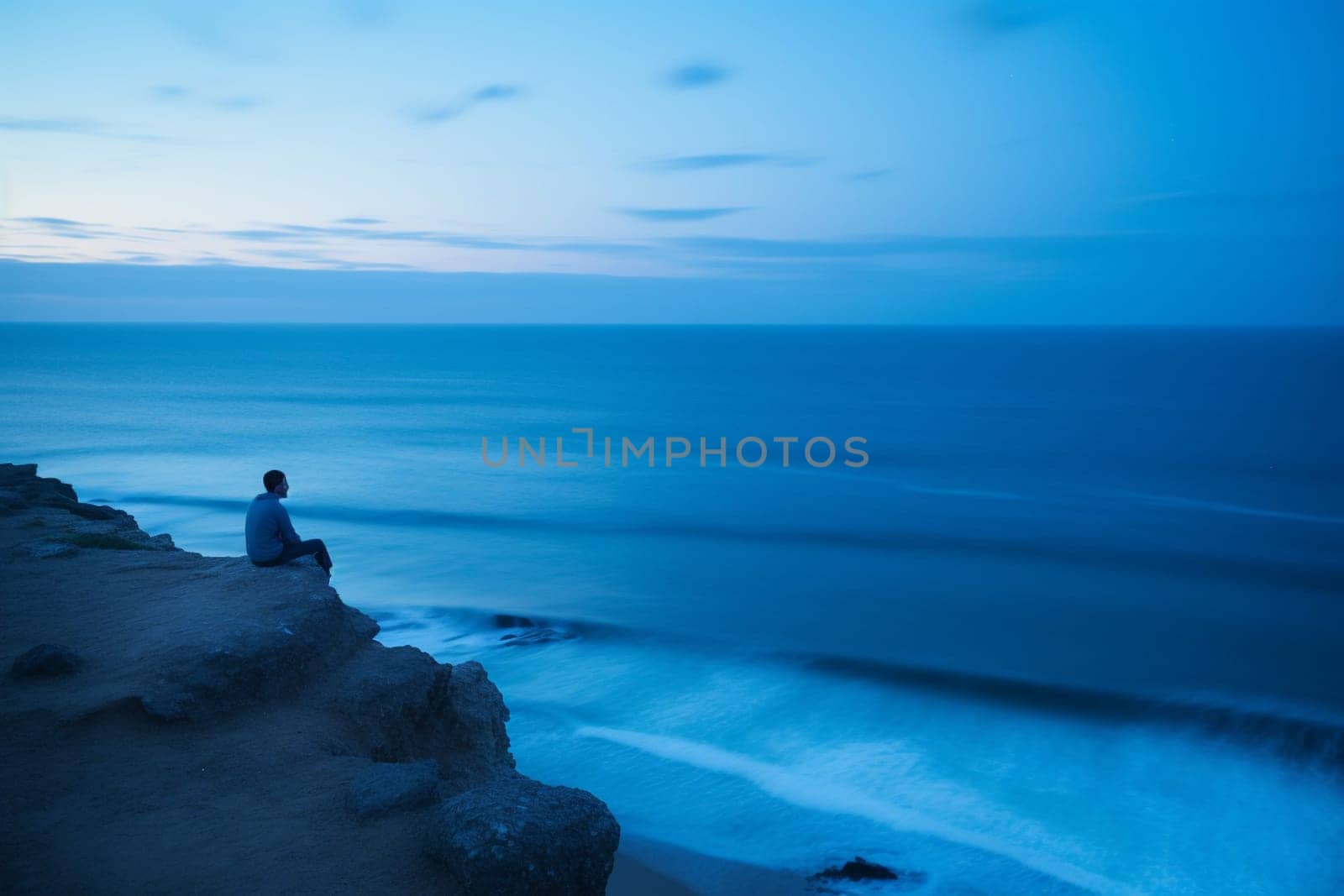 A person sits in meditation at the edge of a cliff overlooking the vast blue of the ocean. The scene is serene and calming, with the blue hour light adding to the reflective and contemplative mood. The sound of the waves crashing against the rocks and the cool breeze help create a harmonious atmosphere for inner reflection and peace. This scene can evoke feelings of calmness, serenity, and introspection. (generative AI)