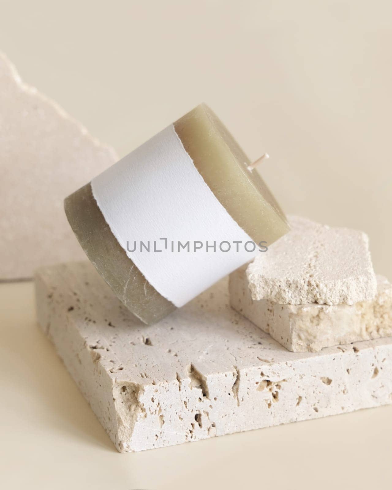 Candle with label on beige near cream travertine stones, Close up, copy space. Brand packaging mock up. Home decoration, minimal interior, Bohemian or scandinavian style