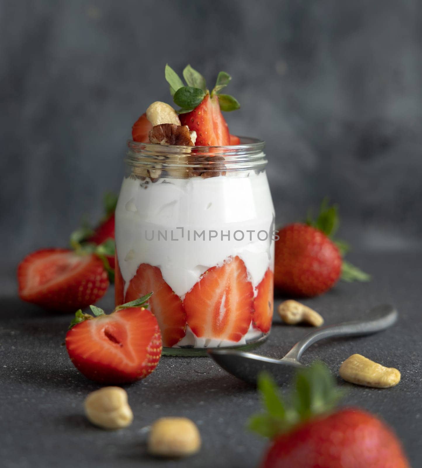 Greek yogurt, nuts and strawberries in a glass jar on grey concrete table with a spoon, close up. Healthy breakfast with fruits and nuts. Great way to eat protein