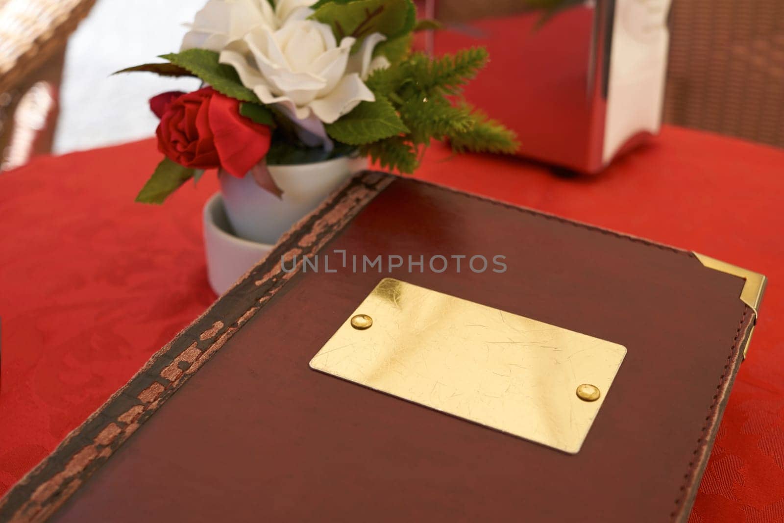 Detail of leather bound menu with blank name plate on red table cloth. There's a small flower vase on the side with red and white roses.