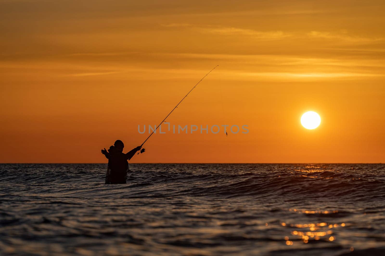The silhouette of an angler in waterproof trousers fishing in front of a dreamy orange sunset in the Baltic Sea. He stands in the middle of the water of the Baltic Sea.