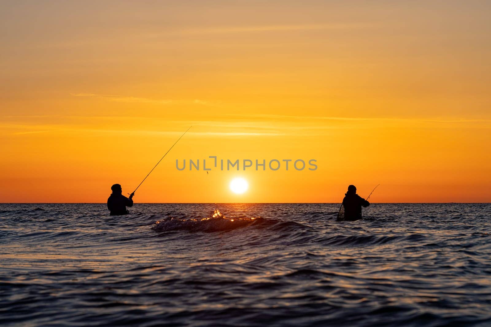 The silhouettes of two anglers in waterproof trousers fishing in front of a dreamy orange sunset in the Baltic Sea. They are standing in the middle of the water of the Baltic Sea.