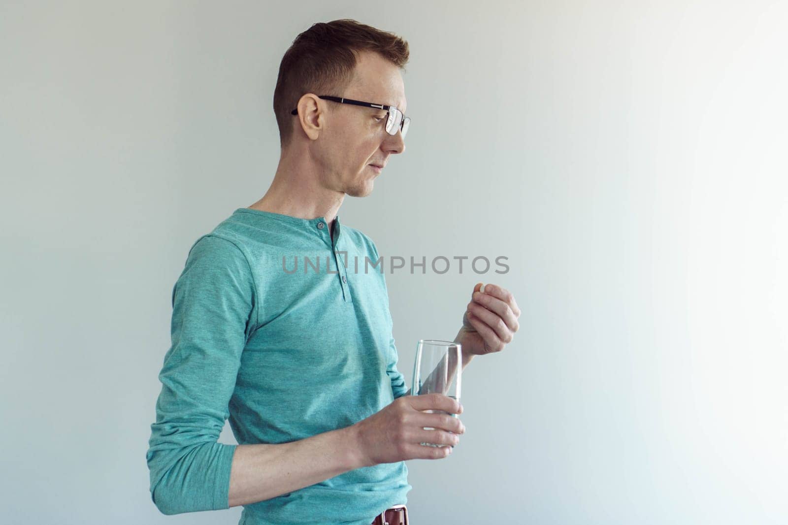 Poor health and stress. Man in glasses and cotton T - shirt stands against light wall with glass of water in his hand and is about to take pill. Painkillers. Antidepressants. Gastroenterology.