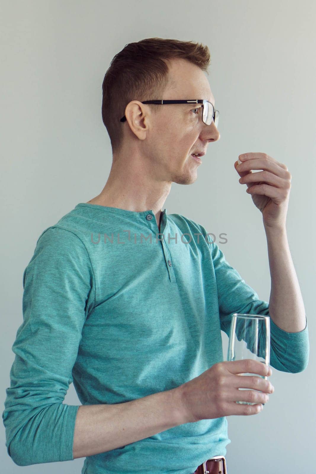 Close-up portrait of man in turquoise cotton T-shirt and glasses, standing on light background with glass of water and drinking pill, vitamins, antidepressants. Painkillers. Gastroenterology.