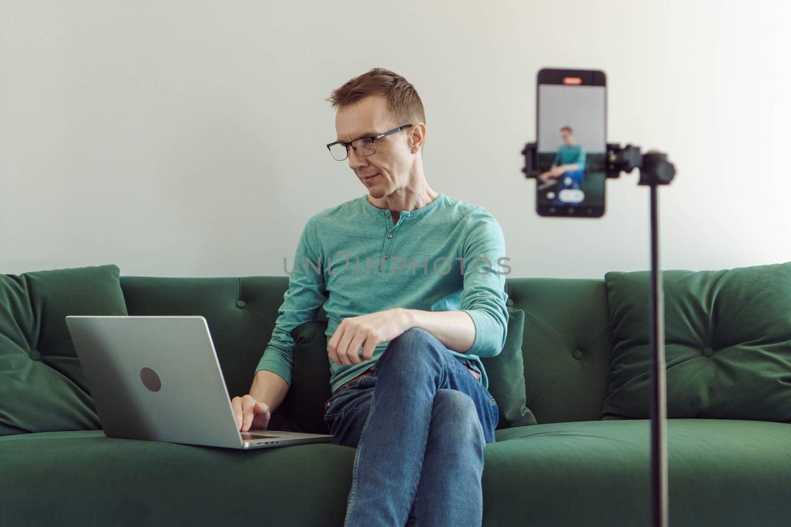 Man in a T-shirt and jeans is sitting at home on couch in front of phone camera and typing something in notebook. Modern technologies. Online training. Male blogger. Video chat.
