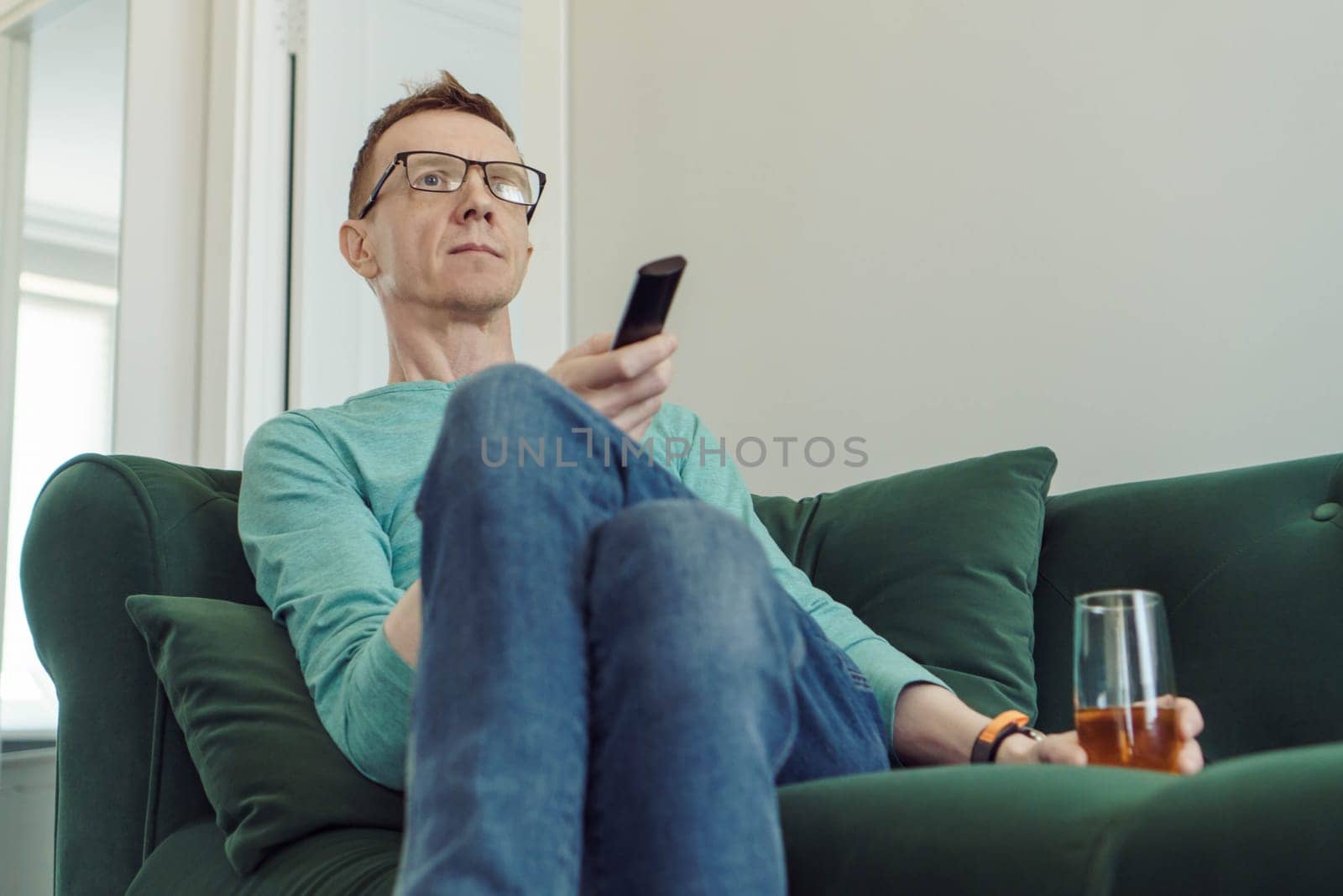 Handsome man in comfortable home clothes is sitting at home on green sofa with TV remote control in his hands and is looking for interesting channel. Free time and rest. Home life. Lonely people.