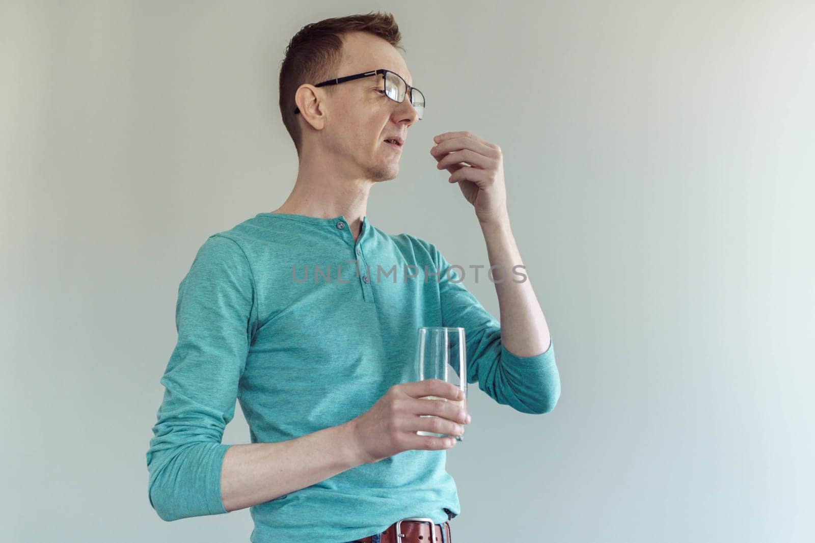 Man in glasses and turquoise cotton T - shirt stands against light wall with glass of water in his hand and is about to take pill. Feeling unwell. Painkillers. Antidepressants. Gastroenterology.