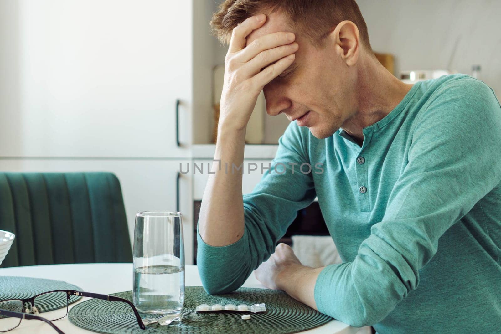 Close-up portrait of man suffering from headache, sitting at home in front of glass of water and pain pills. Illness and loneliness. Medical insurance. Overwork and fatigue. Nervous stress.