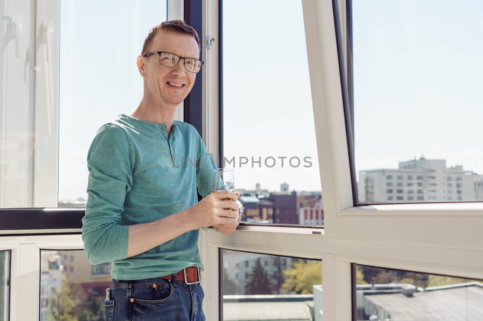 Positive emotions. Man in turquoise T-shirt with long sleeves stands on balcony of house, apartment, office with glass of juice and smiles, looking at camera. City and buildings. Tourism and travel.