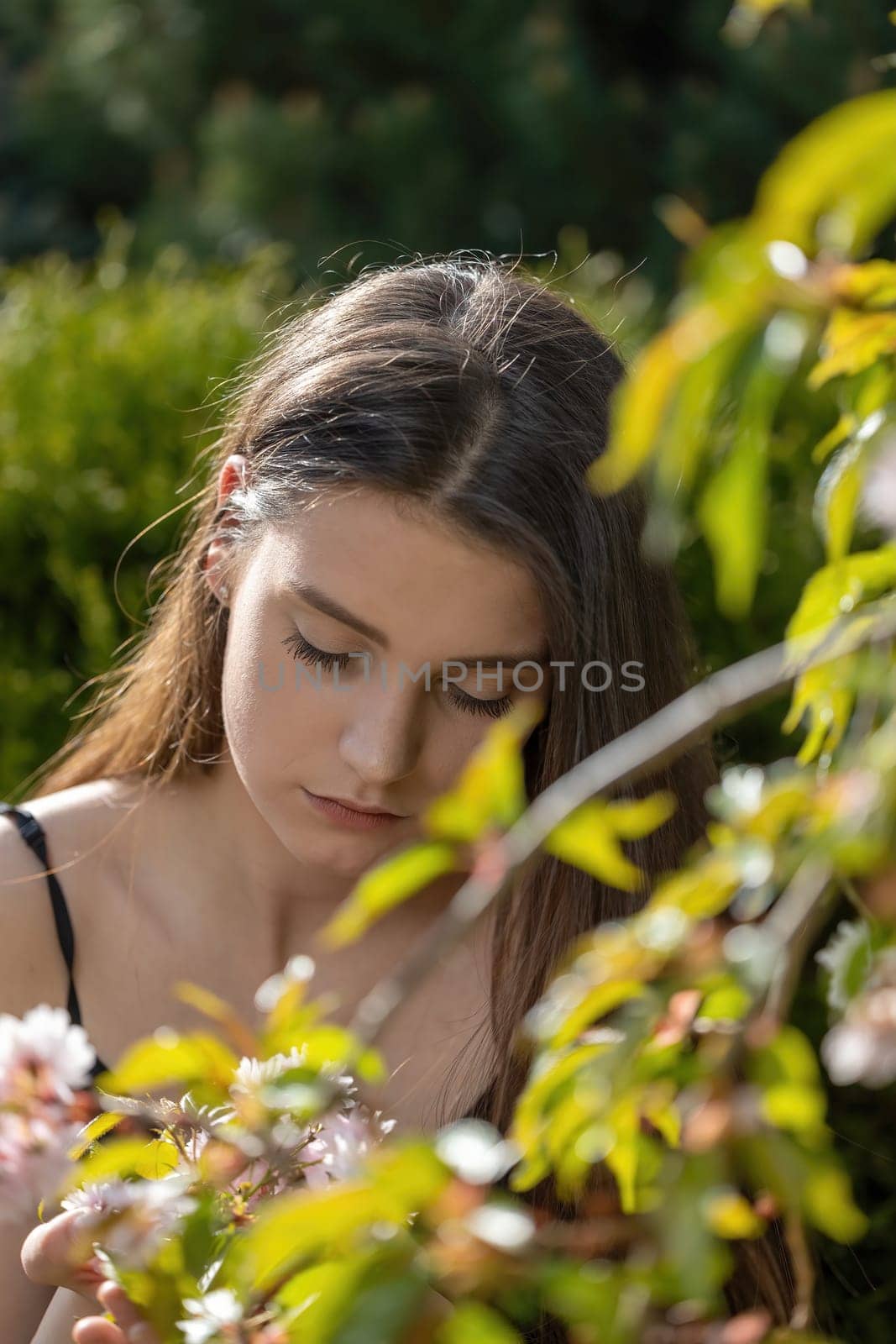 Serious girl poses with downcast eyes behind a blossoming sakura tree. Vertically.