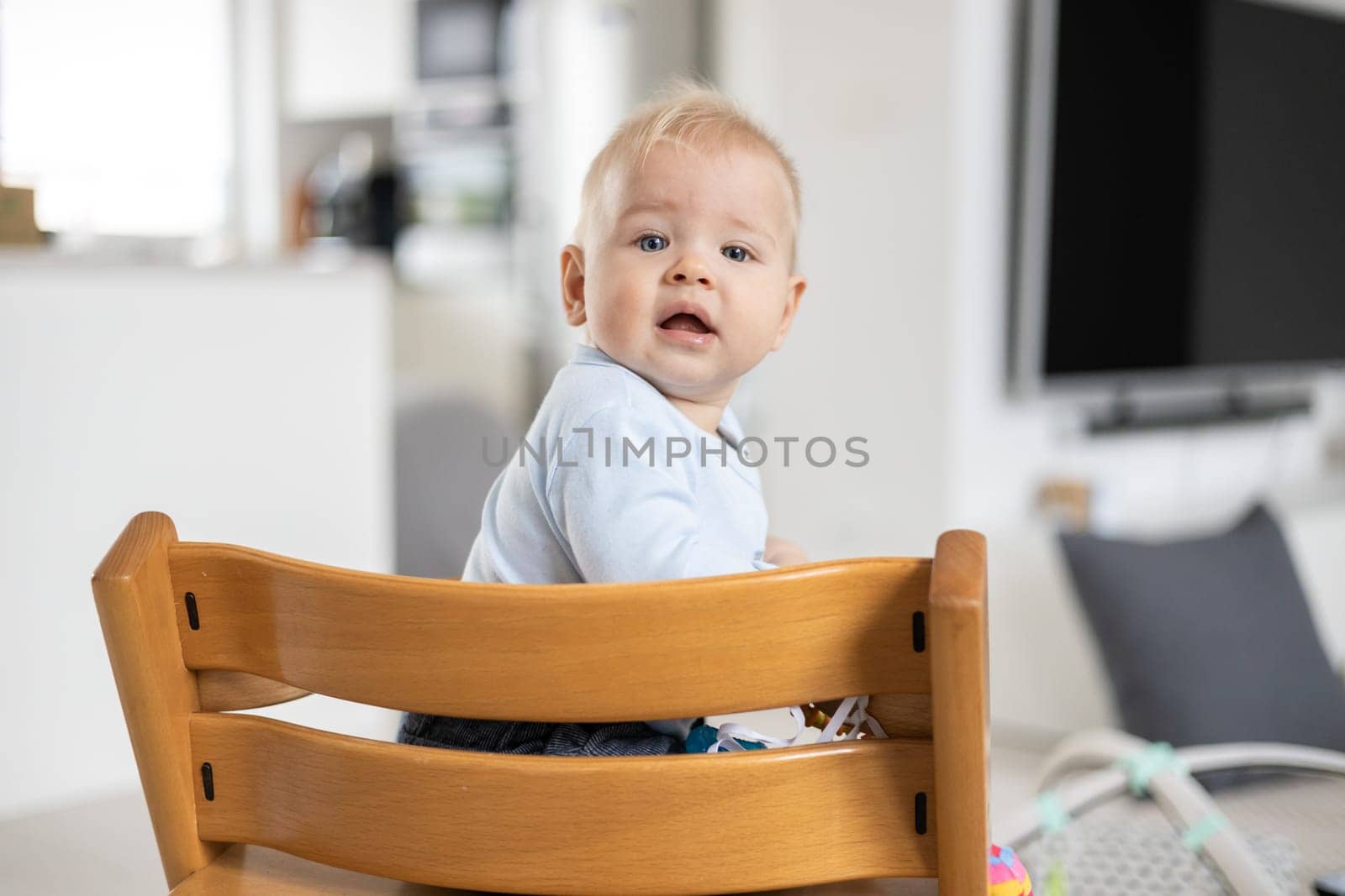 Happy infant sitting in traditional scandinavian designer wooden high chair and laughing out loud in modern bright home. Cute baby smile