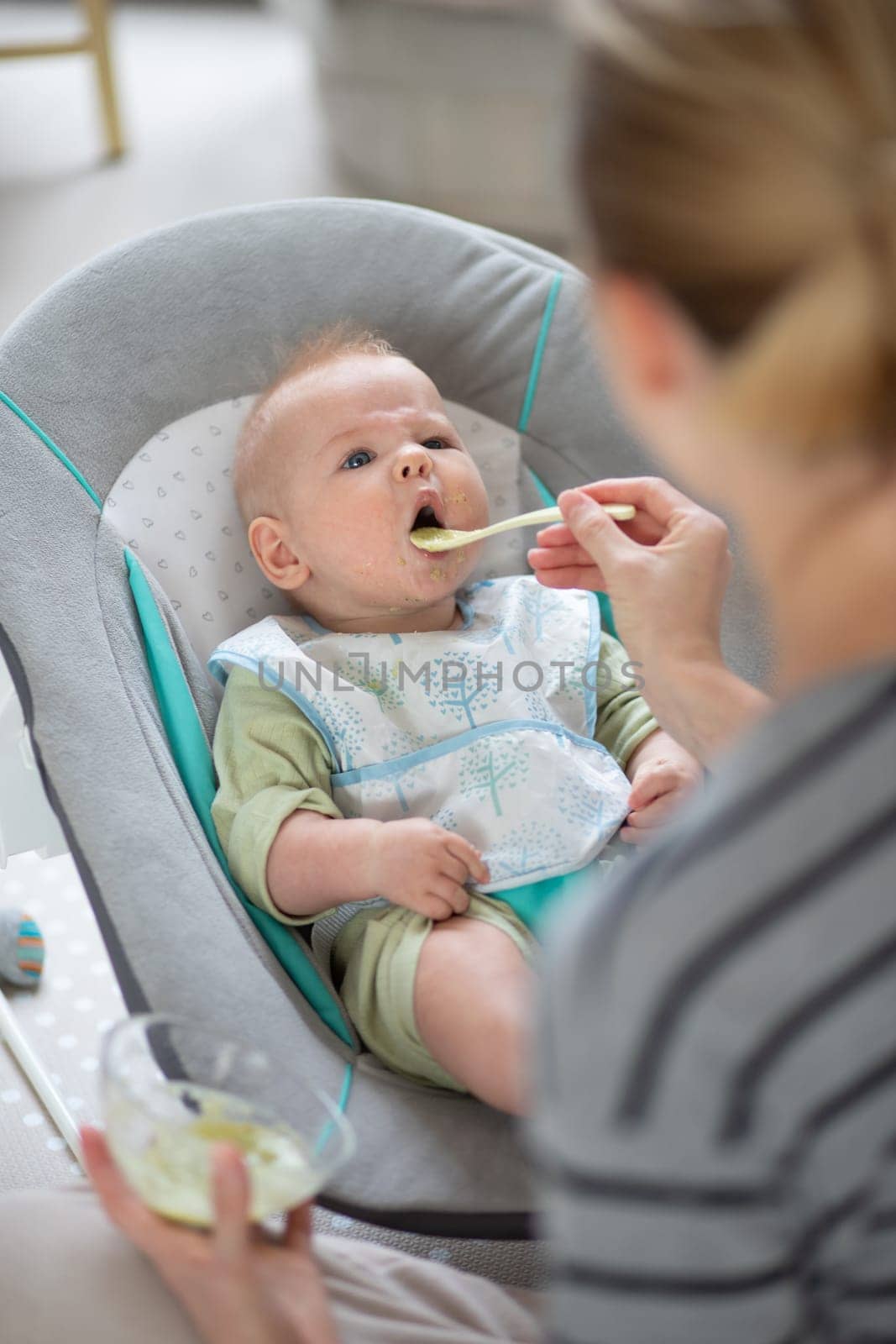 Mother spoon feeding her baby boy child in baby chair with fruit puree. Baby solid food introduction concept