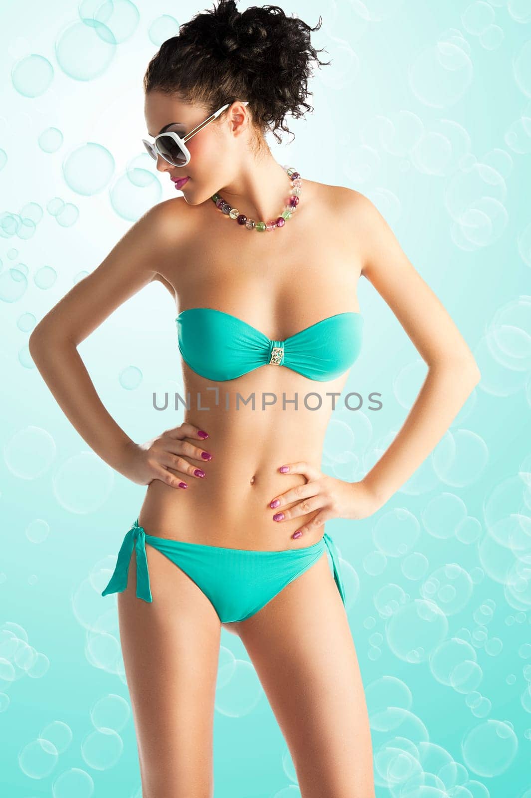 cute sexy brunette in green bikini with white sunglasses and curly hair style over white, she is in front of the camera, her face is turned on profile and she looks down at right. Her hands are both on the hips