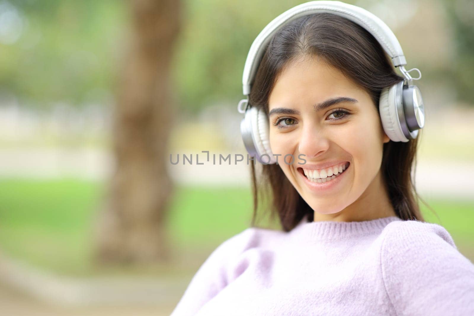 Happy woman with headphone looks at you in a park smiling