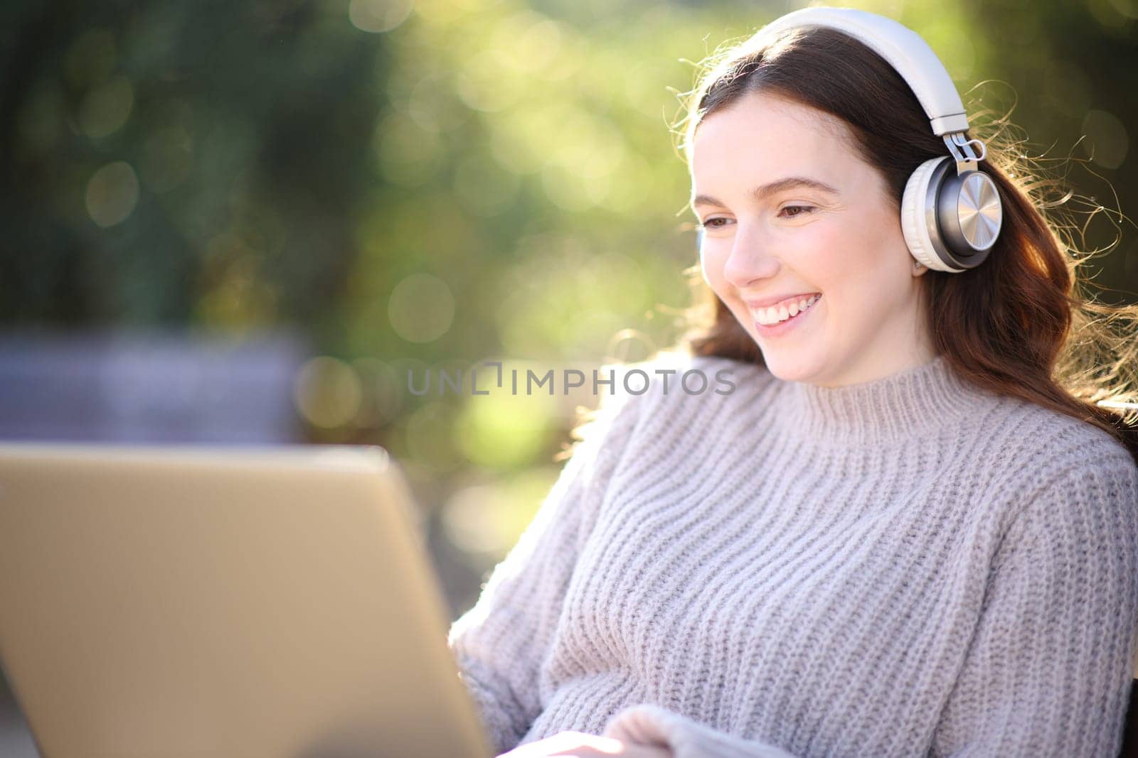 Happy woman listening and watching laptop content sitting in a park with headphone