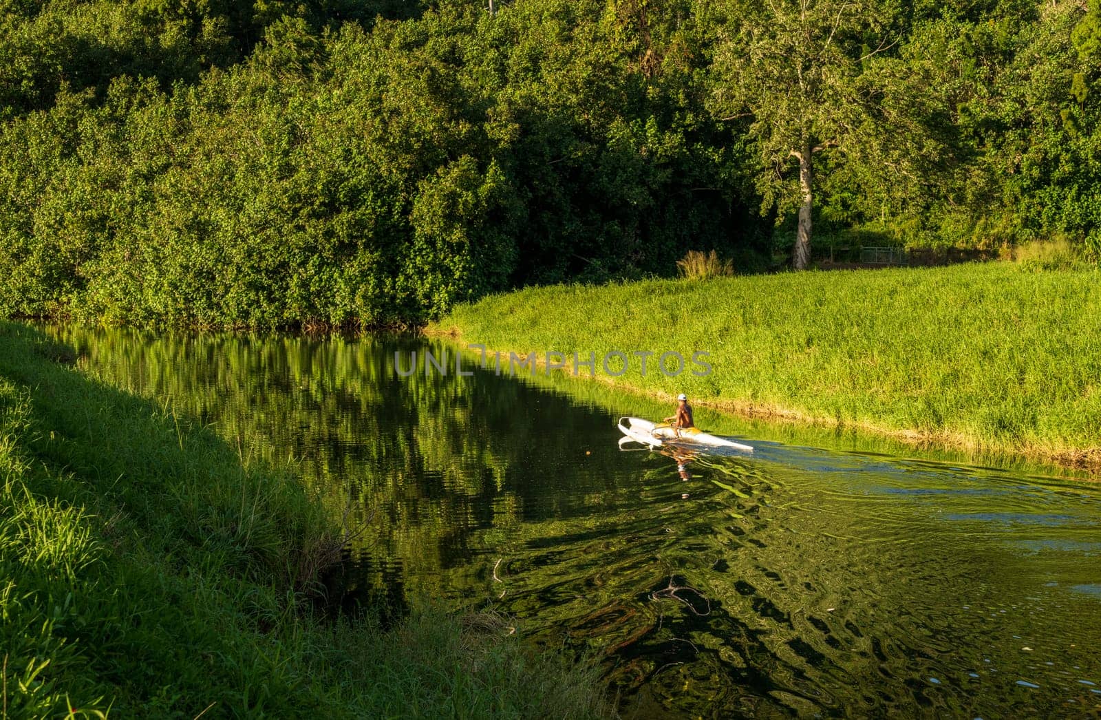 A man paddles a traditional Hawaiian outrigger canoe through the tranquil waters of Hanalei River on a sunny day, creating mirrored reflections.