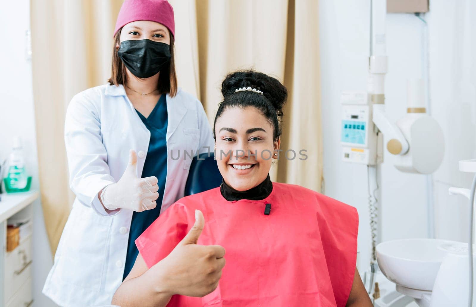 Female dentist with satisfied patient smiling at camera and giving thumb up, Female dentist with patient smiling and giving thumb up. Portrait of female dentist with patient smiling at camera in office