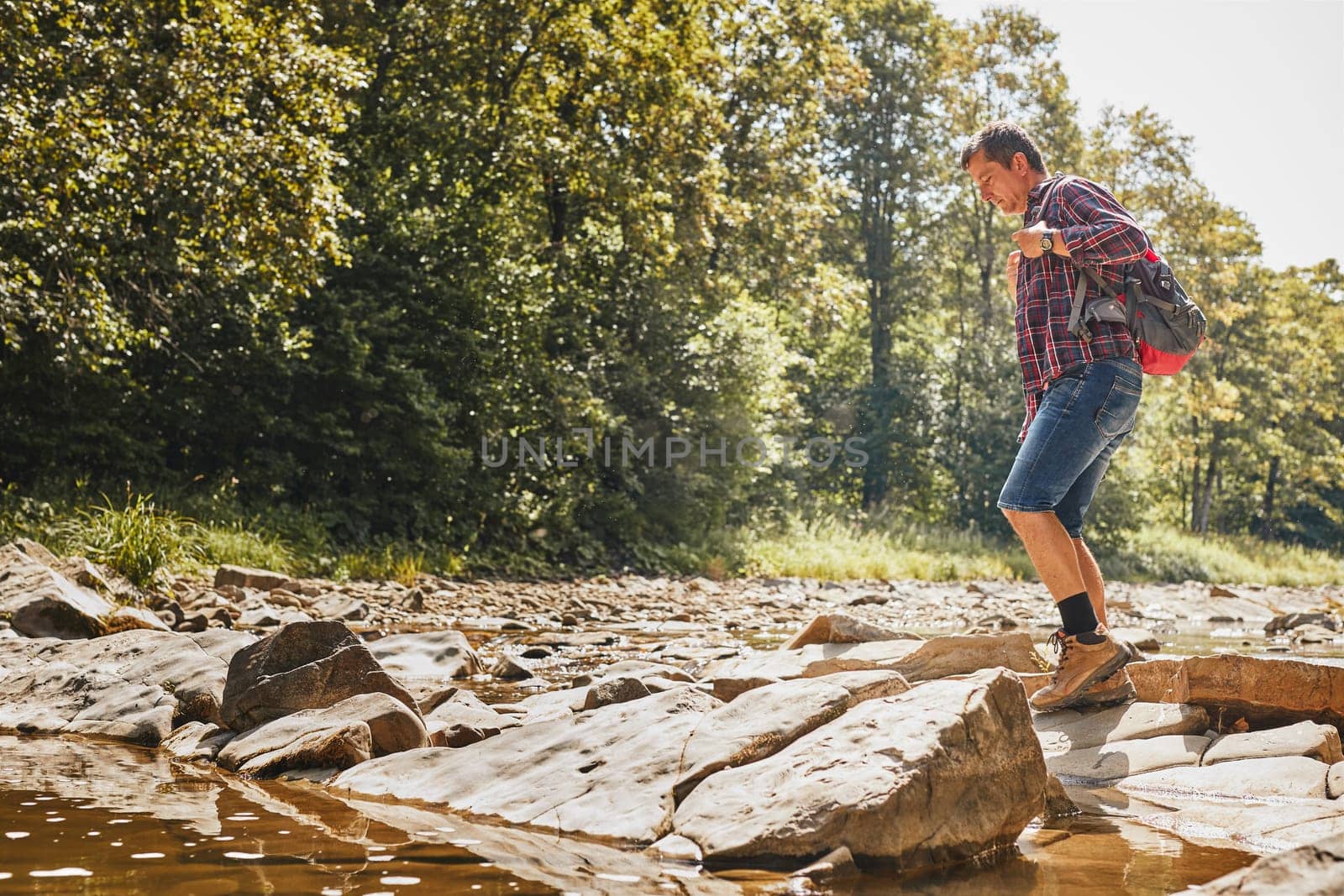 Trekking with backpack concept image. Backpacker wearing trekking boots crossing mountain river. Man hiking in mountains during summer trip. Vacation trip close to nature. Natural scenery