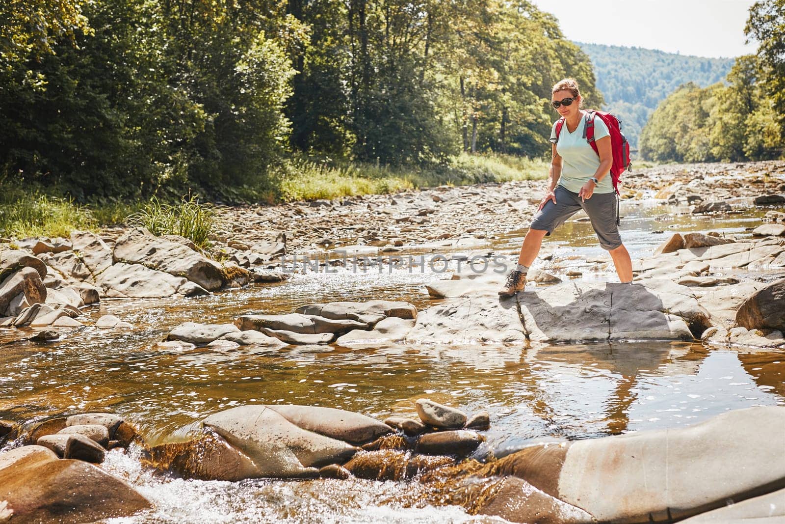 Trekking with backpack concept image. Female backpacker wearing trekking boots crossing mountain river. Woman hiking in mountains during summer trip. Vacation trip close to nature. Natural scenery