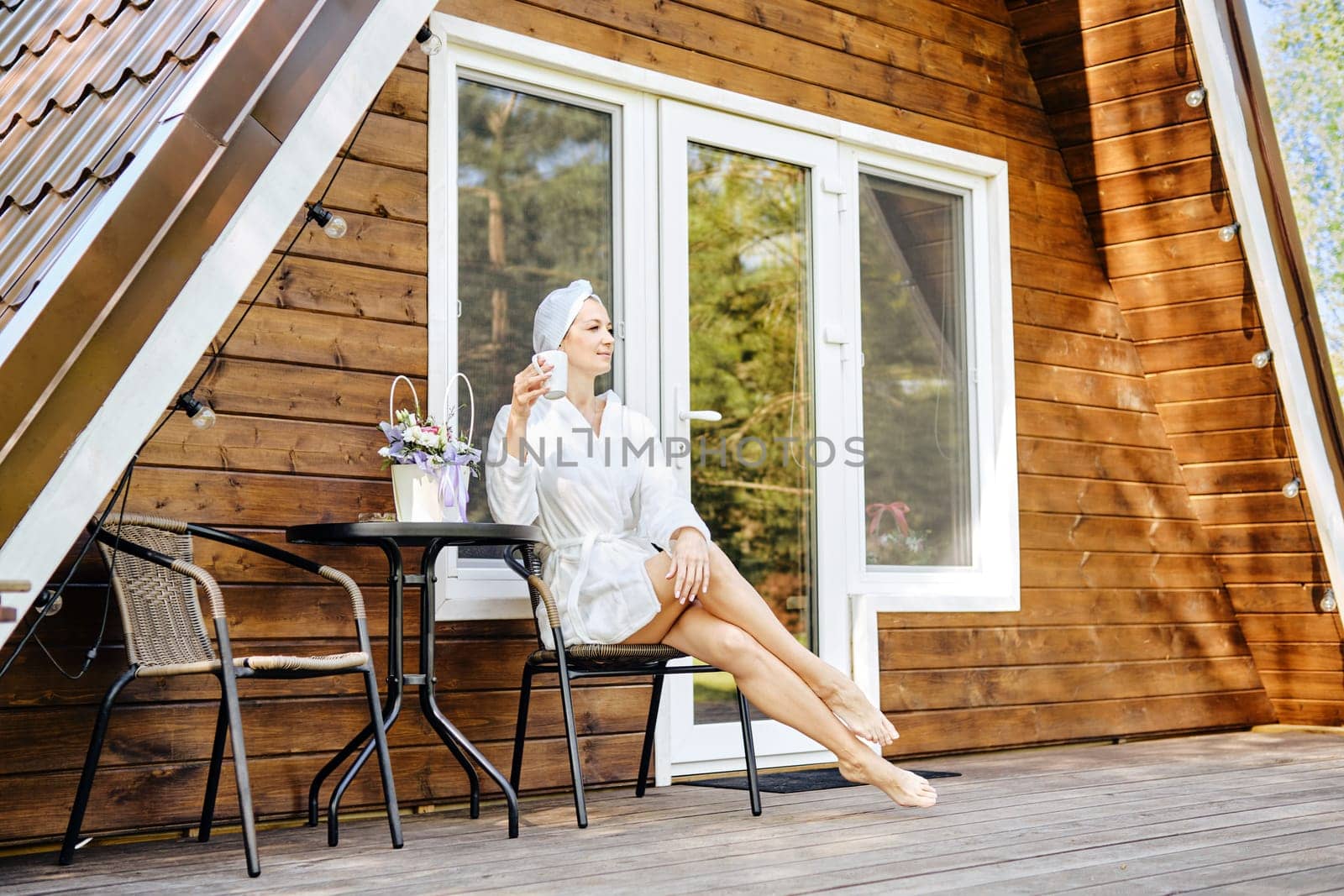 Relaxed woman in bathrobe enjoying morning coffee on terrace of tiny house. Concept of renting of a bungalow to retreat