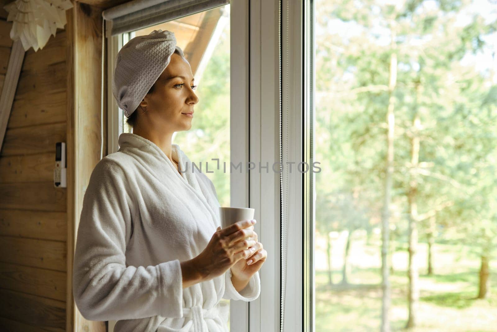 Peaceful woman in bathrobe came to the window of forest house after shower