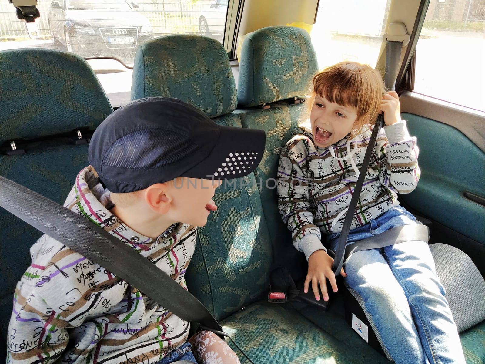Children in the car. A boy and a girl are sitting in the back seat, laughing and grimacing. Children fasten their seat belts. A child in a cap. Children - twins of different faces dressed in the same clothes