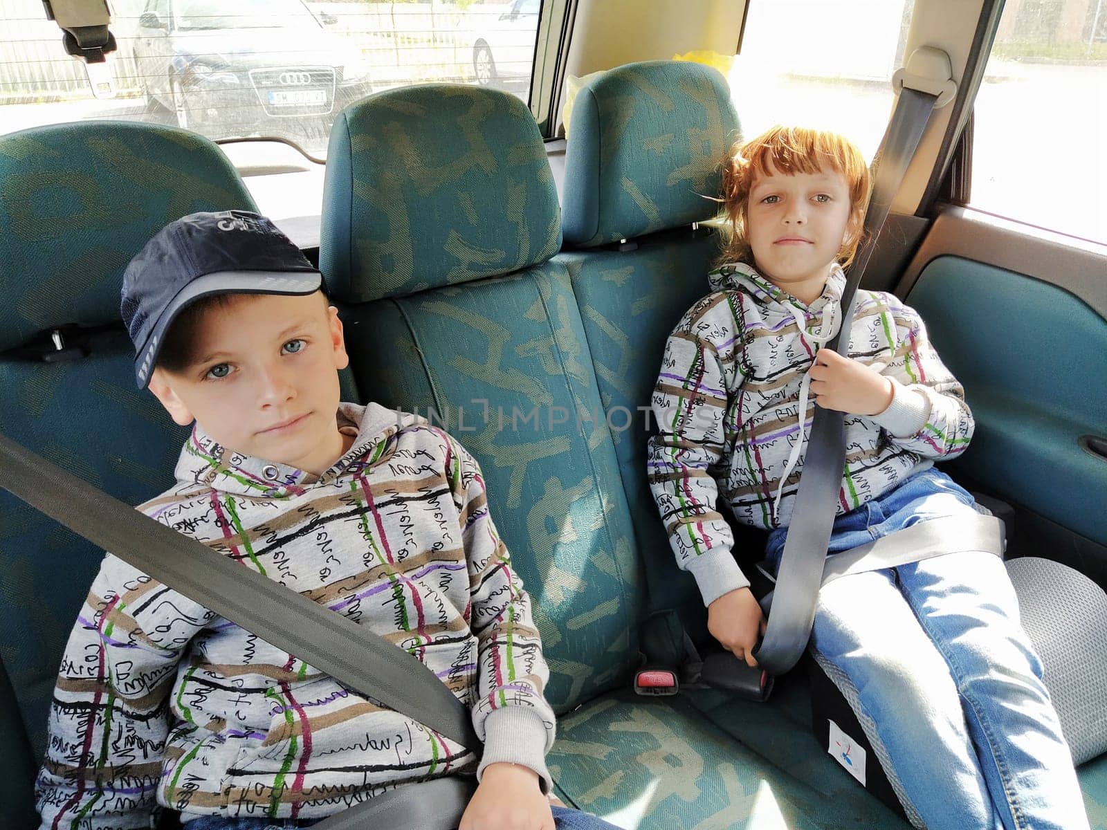 Children in the car. A boy and a girl are sitting in the back seat, laughing and grimacing. Children fasten their seat belts. A child in a cap. Children - twins of different faces dressed in the same clothes