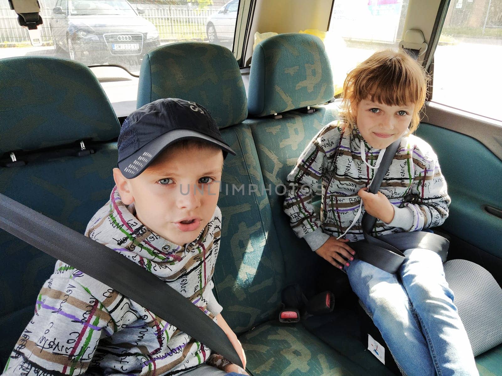 Children in the car. A boy and a girl are sitting in the back seat, laughing and grimacing. Children fasten their seat belts. A child in a cap. Children - twins of different faces dressed in the same clothes