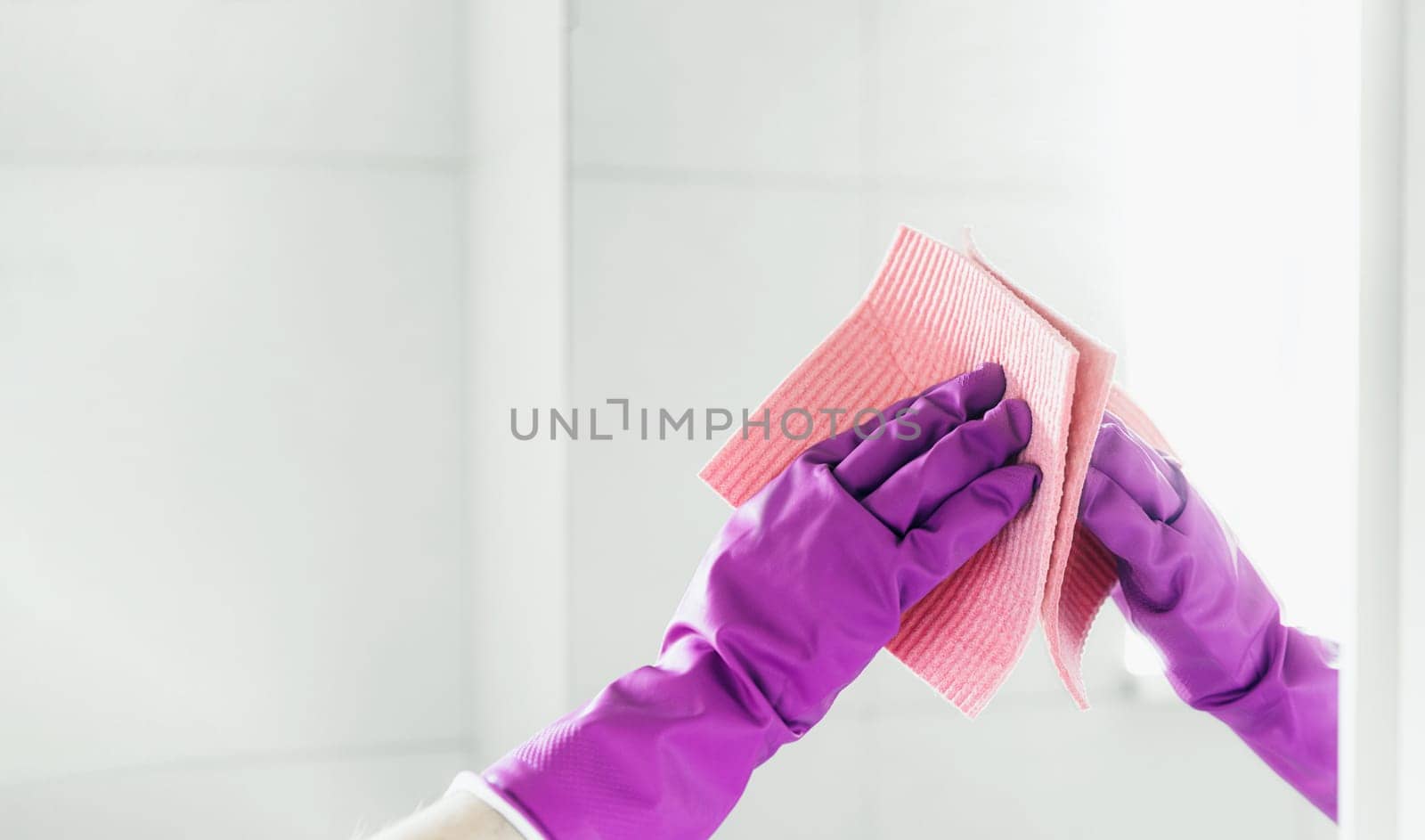 Cropped view of housekeeper woman hand in purple rubber gloves polishing mirror with pink cloth rag cleaner while cleaning bathroom at home. Housekeeping and cleaning service. Clean house, cleanliness