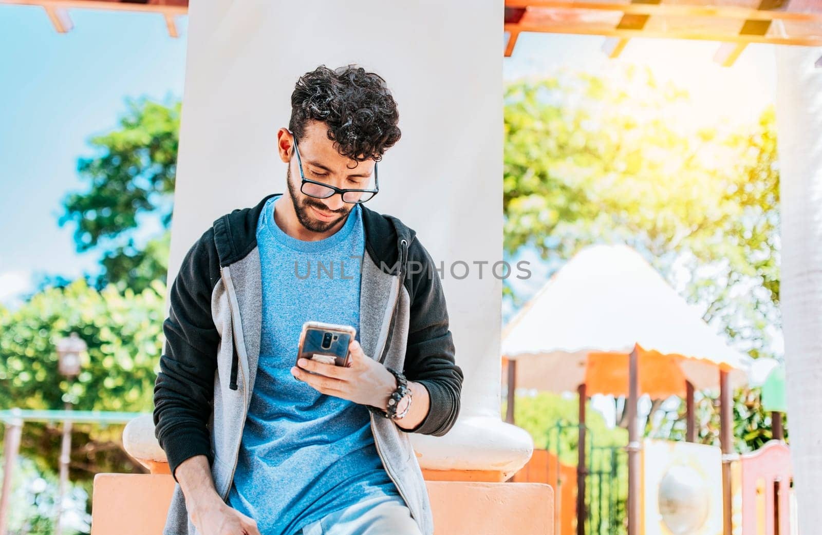 Smiling handsome man using a phone leaning on a wall in a park, Handsome guy in glasses leaning on a wall chatting on his cell phone, Teenage male leaning on a wall sending a text message
