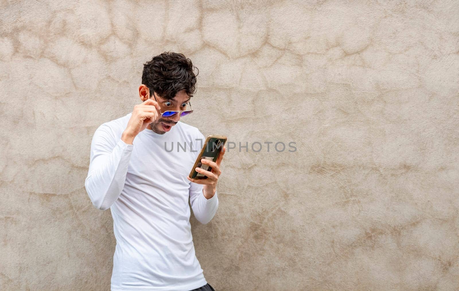 Surprised young Latin looking at his cell phone leaning against a wall. Handsome guy in glasses amazed with his cell phone leaning against a wall, Latin man looking phone leaning against a wall