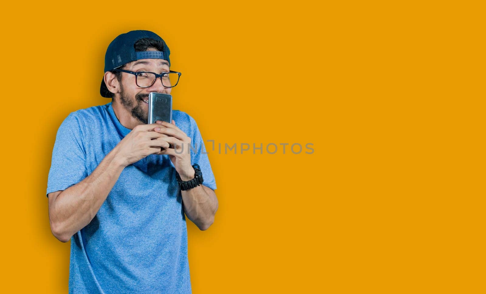 boy in glasses smiling with his cell phone, young man in cap smiling while holding his cell phone on yellow background, boy smiling covering his mouth with cell phone