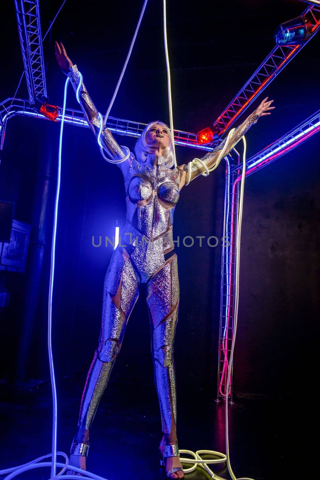 Blonde as a cyber robot. Girl in shining silver armor in neon. Blonde with fantastic makeup and image in dark key and colored light, posing in cyberspace. wires around hands, general plan
