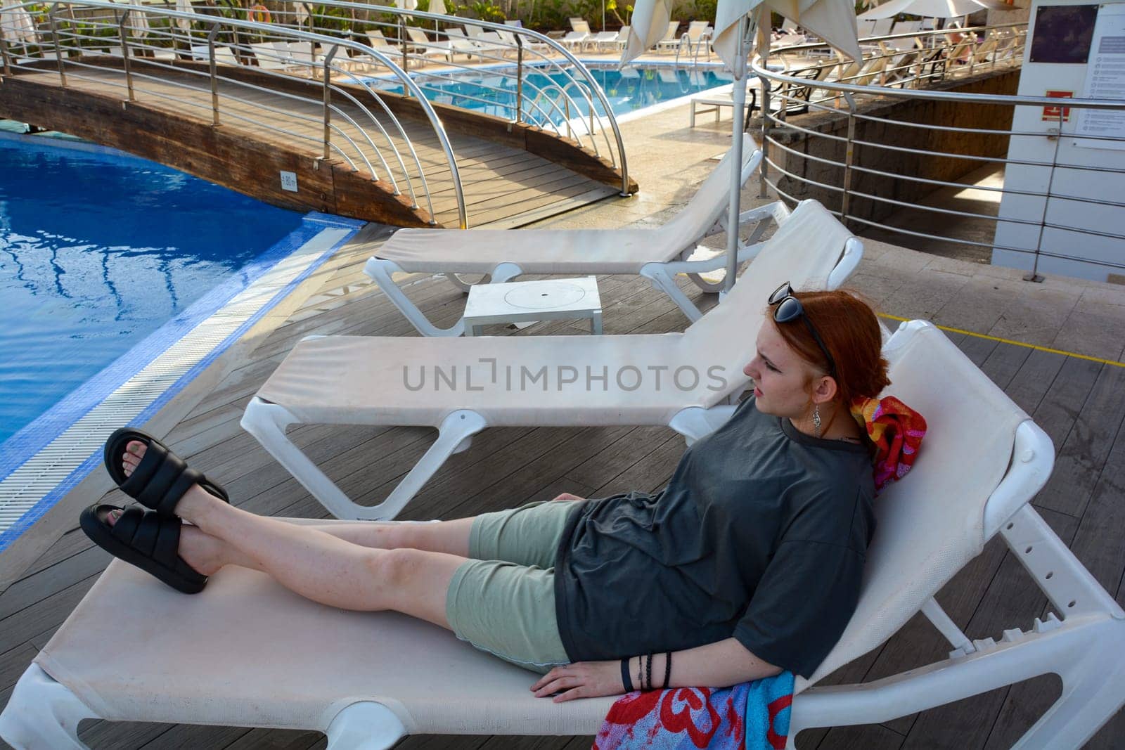 A young teenage girl, dressed in clothes, lies on a sun lounger by the swimming pool