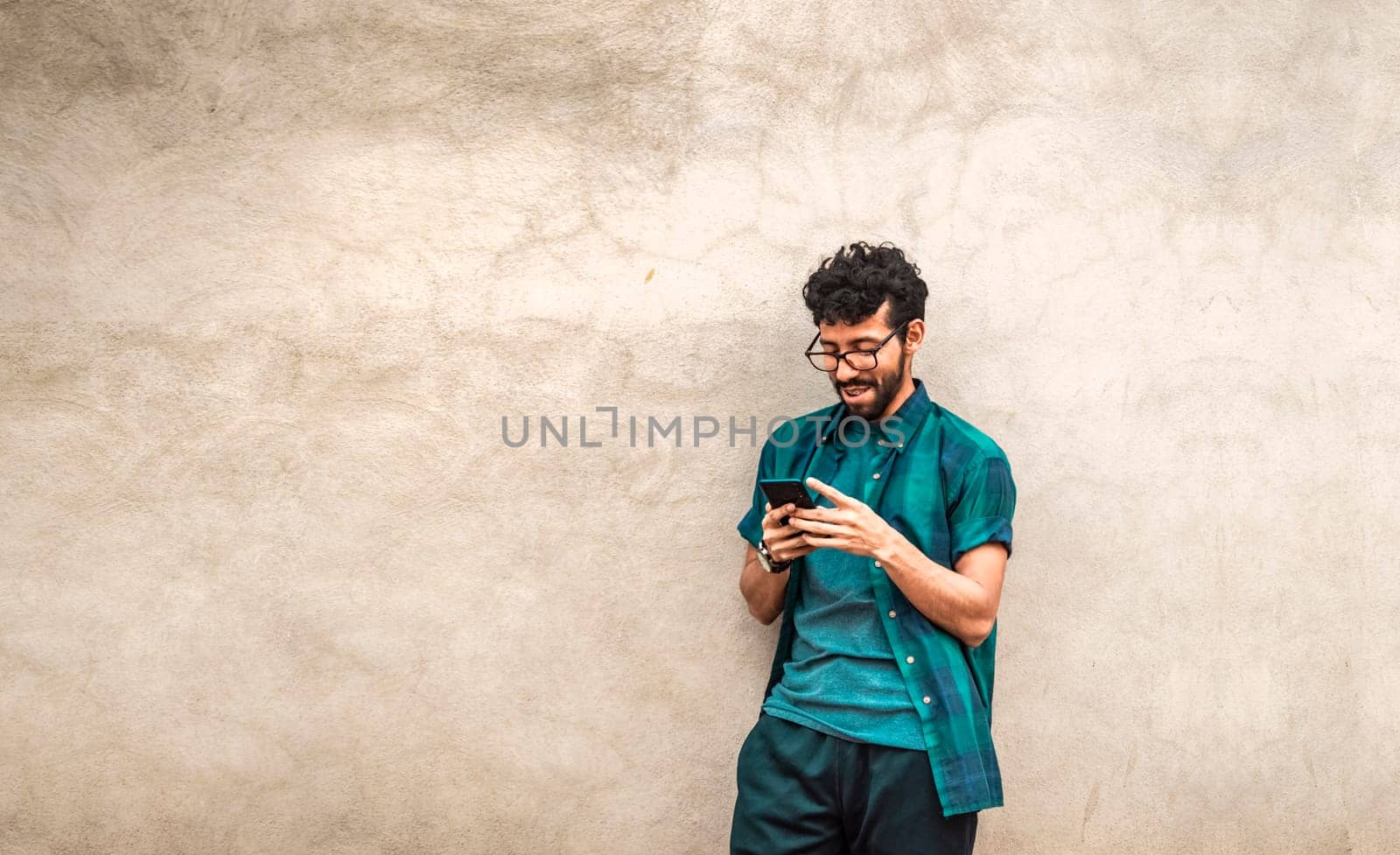 Happy handsome guy leaning against the wall using his cell phone, happy latin man with his cell phone leaning against a wall with copy space, Person smiling phone with space for text