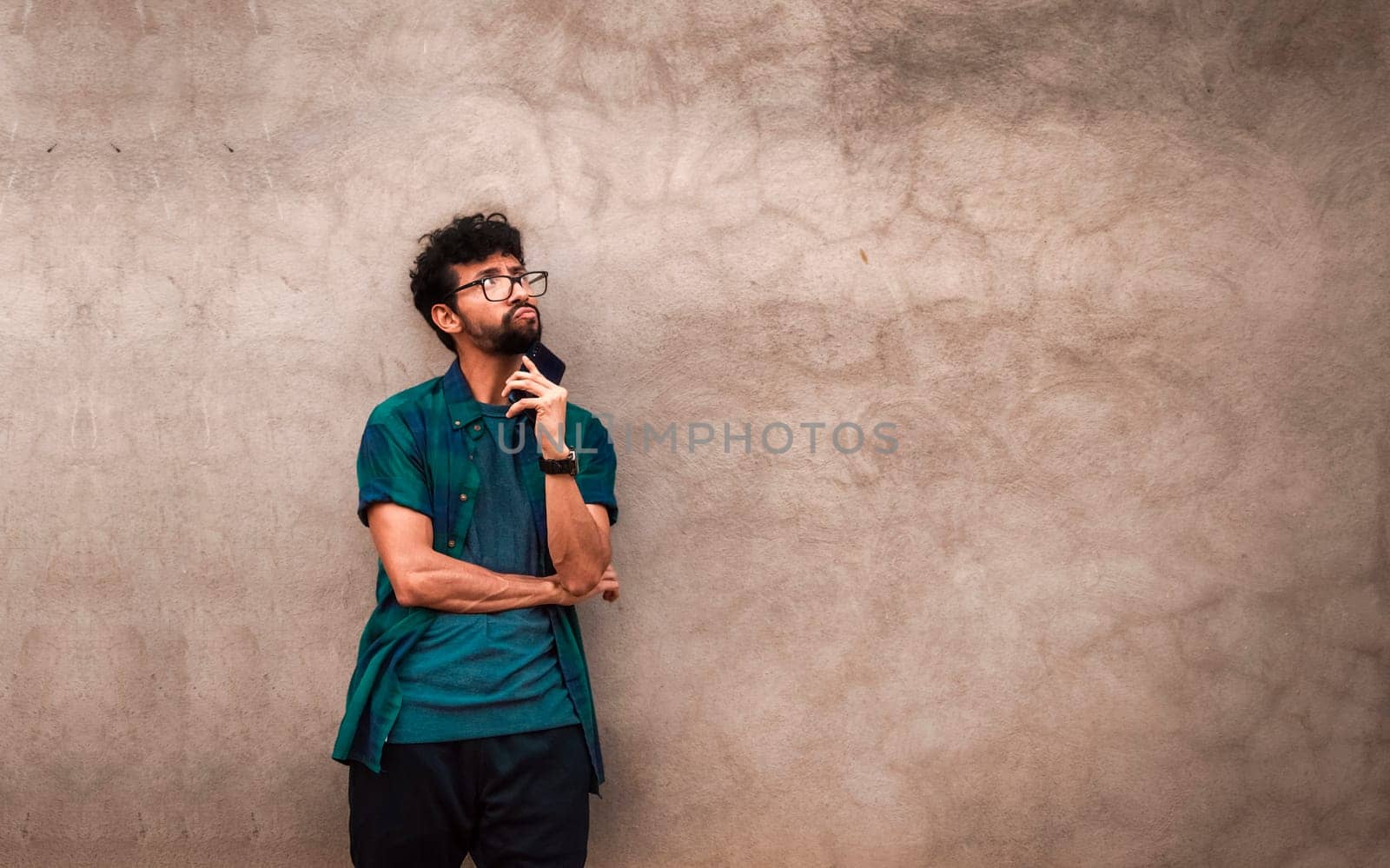 Thoughtful young man with cell phone looking up and touching his chin. pensive latin man with cell phone leaning on a wall, person holding the cell phone pensive and looking up
