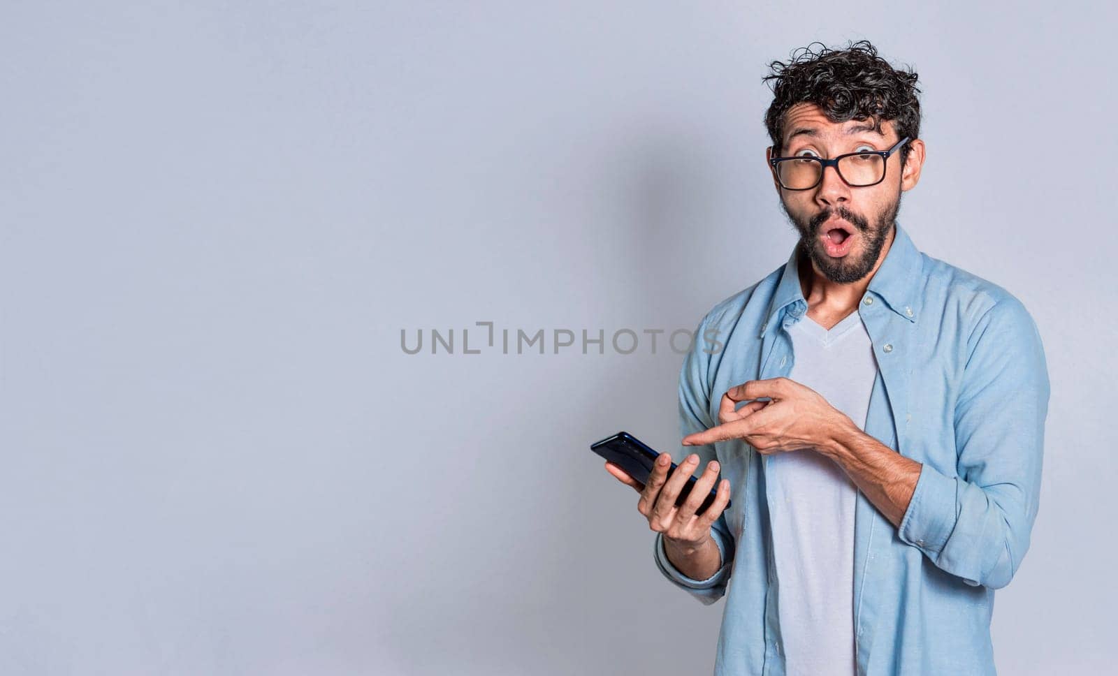 Amazed handsome man pointing at his cell phone isolated, surprised person pointing at his cell phone isolated. Isolated teenager surprised of a promotion on his cell phone