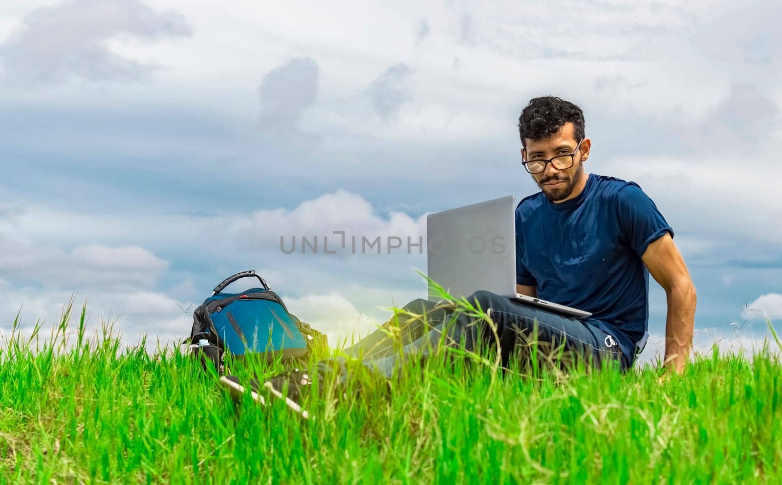 Young man sitting with laptop and backpack in the field. Man sitting in the green field working from his laptop. Freelancer man concept working from the field