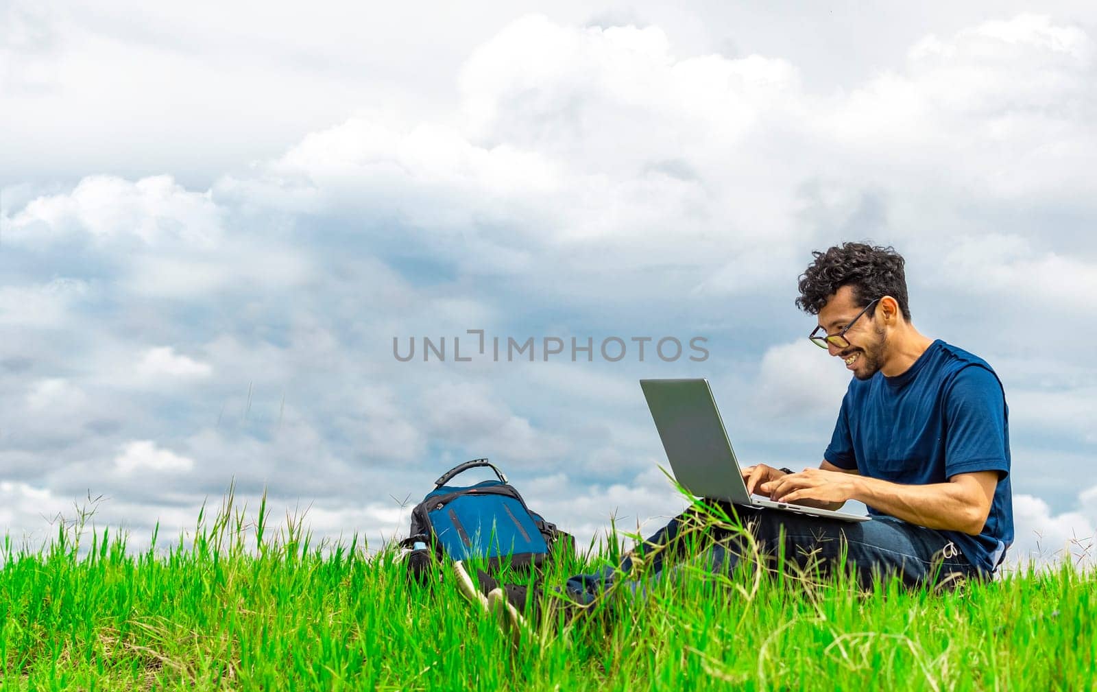 Smiling man with laptop sitting on the grass in the field. Side view of man sitting on green grass working with his laptop. Smiling freelancer guy working from the field