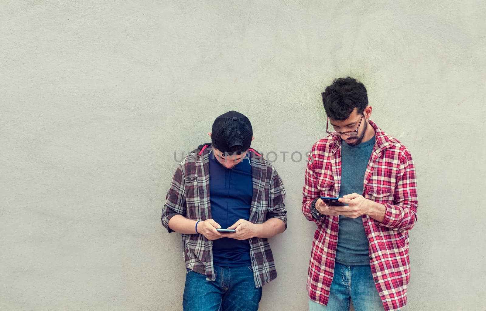 Two teenage friends on a wall checking their cell phones, Two friends leaning on a wall texting on their phones. Friend showing cell phone to his friend, Smiling friends checking cell phones