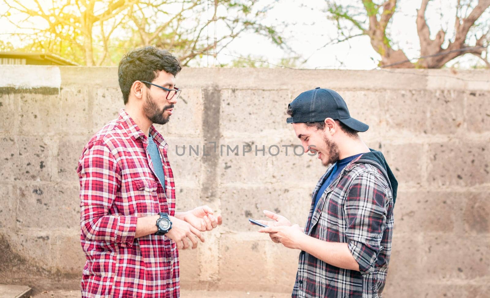 Side view of two young friends chatting outdoors, two friends chatting while smiling and using cellphone. View of two teenagers talking face to face while using the cell phone